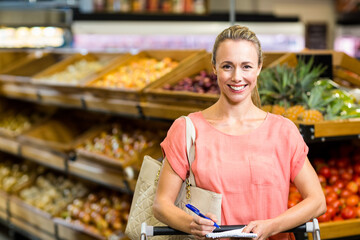 Woman standing in produce aisle holding cart handle noting items on notepad wearing beige bag
