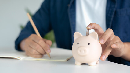 Financial planning and money saving concept with a man's hand putting coins into a piggy bank while writing notes, symbol of investment, budget management, and personal finance.