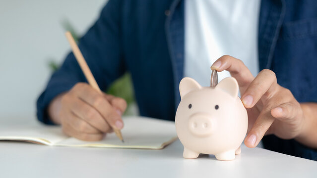 Financial planning and money saving concept with a man's hand putting coins into a piggy bank while writing notes, symbol of investment, budget management, and personal finance.