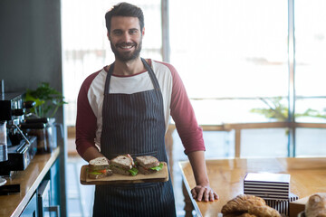 Male barista standing in cafe holding board of lettuce-topped sandwiches beside espresso machine