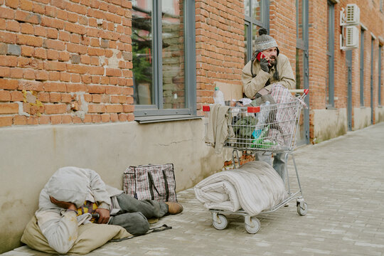 Middle aged Black man lying on sidewalk resting head on pillow near shopping bag, while middle aged Caucasian man standing beside shopping cart talking on smartphone outside brick building