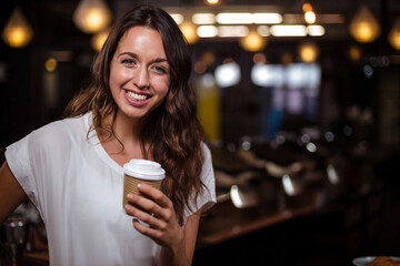 Woman smiling and holding to-go coffee cup inside dimly lit café, with warm pendant lamps overhead