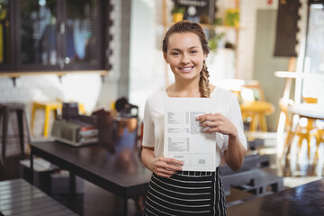 Smiling female server in striped apron holding printed menu at cafe with potted plants