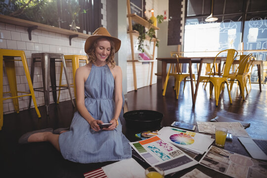 Woman kneeling on café floor, holding smartphone among design layouts, paint brushes, juice