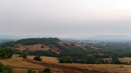 Obraz premium Aerial top view of Vagator hill in Goa during winter, showcasing yellow grass, scattered rocks, green trees, and brown mud paths in a serene landscape.