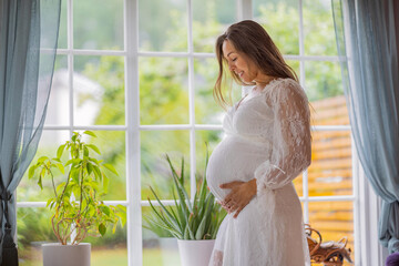 Pregnant woman in white dress by window in a serene home setting
