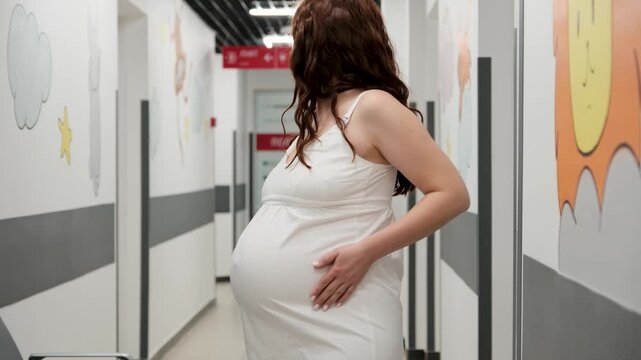 Pregnant woman in white nightie standing in hospital corridor, experiencing labor contractions, gently touching her belly while waiting for childbirth, embodying anticipation of motherhood