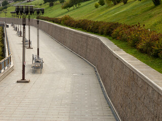 Perspective view of a long, curving promenade in a tranquil city park. A stone retaining wall and a...