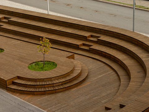 A high-angle view of a modern public amphitheater, showcasing contemporary landscape architecture. The curved, tiered structure is crafted from warm brown wooden decking, with a young tree growing fro