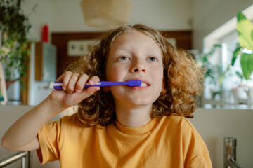 A child with curly hair smiles while brushing their teeth in a bright bathroom. Natural light filters in, enhancing the cheerful atmosphere of morning hygiene.