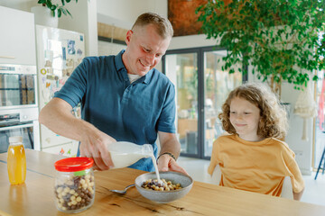 A father smiles as he pours milk into a bowl of cereal for his son in a bright, modern kitchen. The child watches happily, creating a warm moment shared over breakfast.