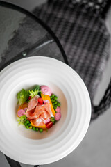 Gourmet appetizer with cured meat, peas, cherry tomatoes, pink sauce, and greens on a white plate. Artistic restaurant presentation, top view on glass table.