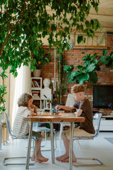 Two boys are seated at a wooden table in a bright room, surrounded by plants. They are playing a board game, enjoying quality time together while their father is nearby.