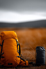 Orange hiking backpack on mountain trail with blurred landscape, outdoor camping gear in wilderness, adventure travel equipment on rocky terrain, trekking rucksack in nature setting
