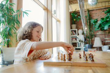 A young boy with curly hair smiles as he plays chess alone at a wooden table in a cozy room filled with plants. Sunlight filters through large windows, creating a warm ambiance.
