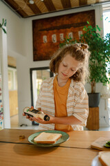 A young boy adds seasoning to a slice of bread while standing in a well-lit kitchen. His focused expression reflects his engagement in the cooking task alongside his family.