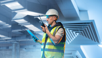 HVAC supervision. Worker in safety vest and helmet inspects ventilation ducts, using a tablet and walkie-talkie to coordinate air conditioning and building climate control installation.