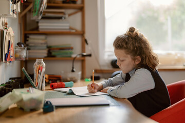 A young boy sits at a wooden desk, focused on writing in his notebook. Various art supplies and...