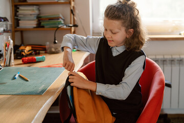 A young boy sits in a red chair, packing his backpack on a wooden desk. He focuses on arranging his school supplies, surrounded by books and art materials during morning preparations.