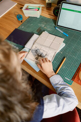 A boy sits at a wooden desk engaged in drawing. He interacts with his notebook and art supplies while a tablet displays various information next to him. The room is well-lit and organized.