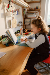 A young boy engages in creative activities at his desk, surrounded by books and craft supplies. He writes with a pencil, showcasing his imagination and concentration while enjoying time at home.