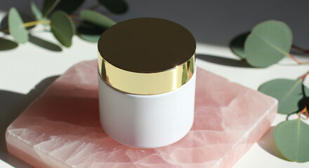 Photo of a luxurious white cosmetic jar with a gold lid rests on a pink rose quartz stone, adorned with eucalyptus leaves