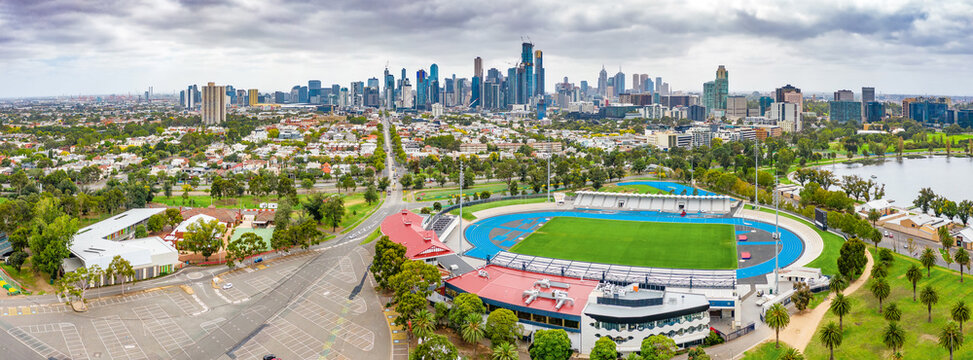 Aerial panorama of a sports arena and carpark in parkland in front of a city skyline at Albert Park