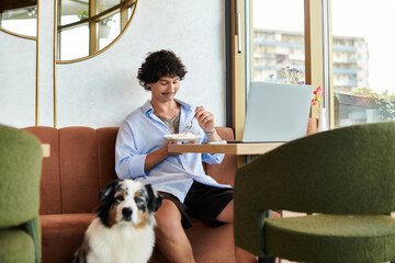 Young man enjoys lunch while working on laptop in a trendy cafe with his dog by his side