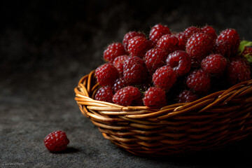 Fresh red raspberries in rustic wicker basket on dark black background, ripe organic berries with dramatic moody lighting, healthy antioxidant fruit for dessert snack, natural sweet summer harvest 