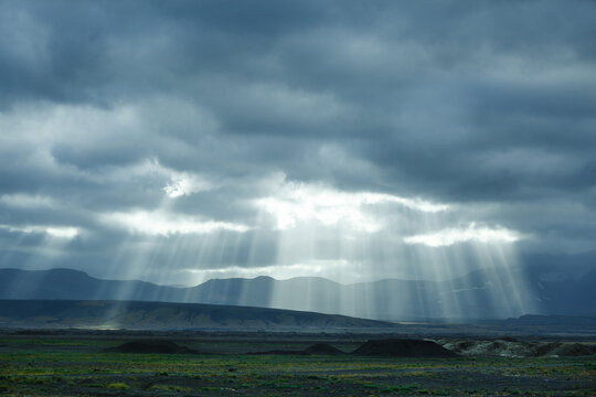 Dramatic landscape of Iceland. Gloomy weather with sunbeams breaking through heavy clouds and falling on waste land barely covered with short green grass and low mountains seen on the background.