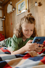 A young boy lies on a vibrant bedspread, focused on his smartphone. He is smiling as he interacts with the device, creating a moment of leisure in his cozy room.