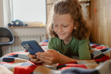 A young boy lies comfortably on his bed, focused on his smartphone. Soft light filters through the window, creating a cozy atmosphere as he enjoys his time alone.