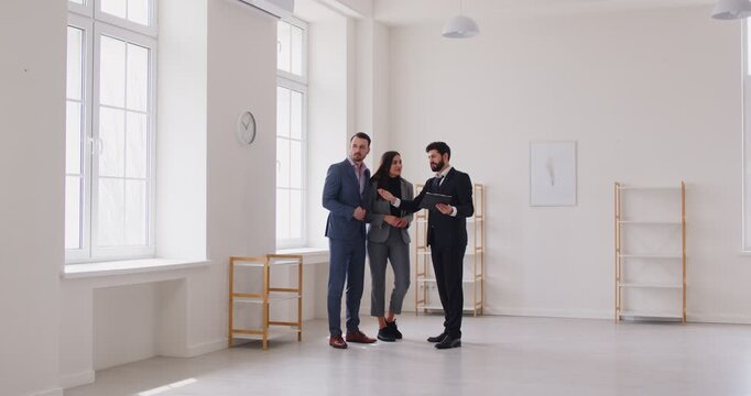 Young couple visiting empty apartment with male realtor during property viewing. Real estate agent describing features of modern flat to clients and discussing possible conditions of purchase or rent.