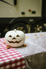 White jack-o'-lantern sits on picnic table with tablecloths at campsite near camper van with copy space, October and camping concept 