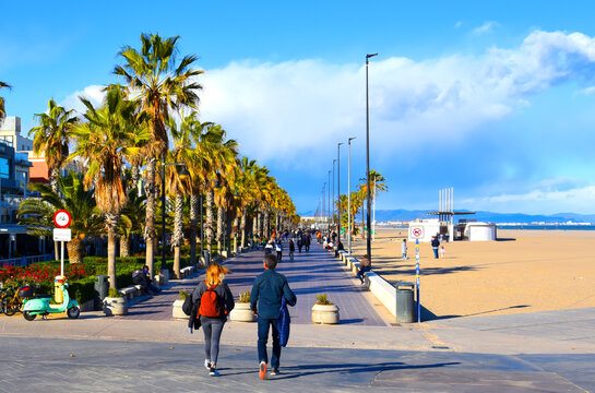 People  walk on path at coastline in autumn holiday season. Winter season in Spain. Couple walking at seafront with Palm trees of La Marina de Valencia. Tourists stroll  coastline on weekend in Spain. - Powered by Adobe