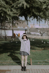 Teen girl in a gray cardigan and plaid skirt standing outdoors, enjoying the sunny day.