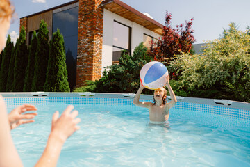 A young boy plays in a pool on a sunny day, tossing a colorful beach ball. His brother is nearby, ready to catch it, while their home is visible in the background.