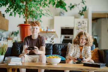 Two boys sit on a comfortable couch, engrossed in playing video games. They share a light moment, surrounded by a bright living space with plants and books.