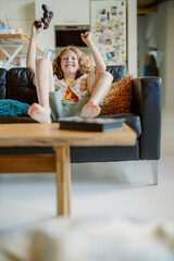 A young boy sits on a sofa, raising his arms in excitement while holding a game controller. He enjoys playing video games in a cozy living room with family nearby during the day.