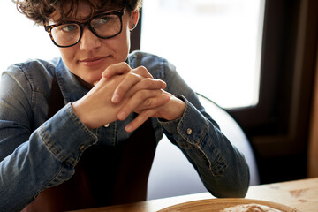 Portrait of confident female entrepreneur with short curly hair and eyeglasses, sitting at wooden counter in her store, thinking over something and smiling slightly, her clasped hands touching chin.