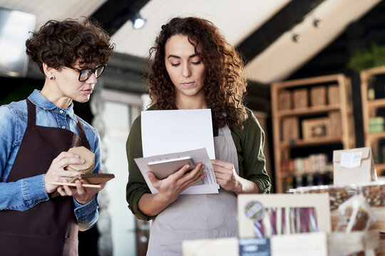 Two concentrated female entrepreneurs in aprons working in a store with natural goods. Woman with long curly hair looking through documents, her colleague helping her, standing beside with notepad.