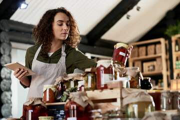 Beautiful curly shop assistant in apron working in small store with organic goods. She standing near homemade jars with pickles and jams, setting them up on wooden boxes and checking its quality.