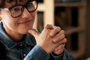 Extreme close-up portrait of pensive entrepreneur in denim shirt and apron at work. Attractive woman with short curly hair and glasses, sitting with her fingers on chin and smiling dreamily.