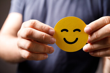 Close-up image of hands holding a yellow smiley face, representing happiness, positivity, and good...