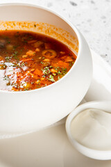 Hot vegetable soup with herbs, tomatoes, and olives served in a white bowl with sour cream on the side. Rustic presentation on a speckled countertop. Top view, natural light.