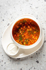 Hot vegetable soup with herbs, tomatoes, and olives served in a white bowl with sour cream on the side. Rustic presentation on a speckled countertop. Top view, natural light.