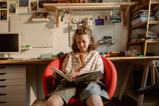 A young boy sits comfortably in a red chair, engrossed in a book. His room is decorated with personal items, showcasing a creative environment filled with memories and activities.