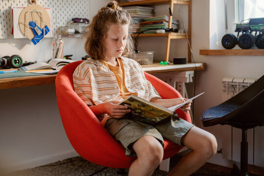 A boy sits comfortably in a red chair, reading a book in his study during the afternoon. Nearby, his workspace features various toys and books, hinting at family time and creativity.