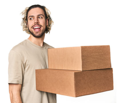 Young man with cardboard boxes in studio looks aside smiling, cheerful and pleasant.