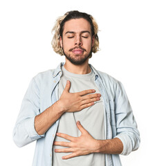Young Caucasian man in studio setting laughs happily and has fun keeping hands on stomach.